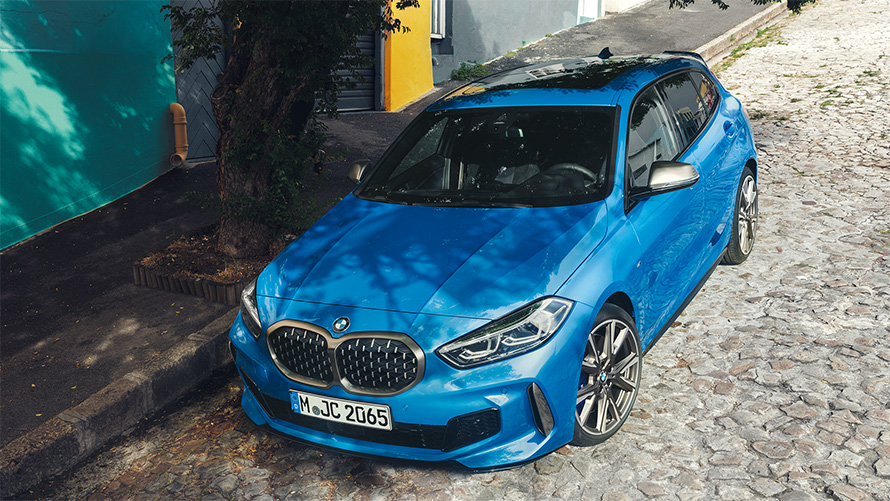 La BMW Série 1 M135i F40 en Misano Blau metallic, vue de trois quarts avant en plongée, au bord de la route en stationnement.