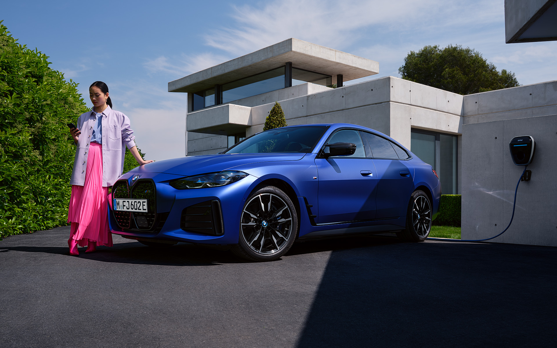 BMW i4 M50 G26 2021 BMW Individual Frozen Portimao Blau metallic, vue de trois quarts avant avec femme devant une maison