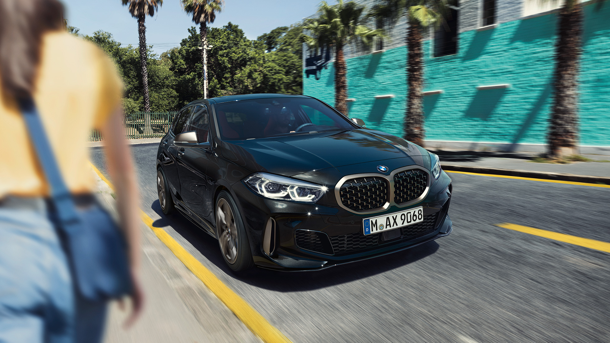 BMW M135i xDrive, vue de trois quarts avant. BMW M135i xDrive en Saphirschwarz metallic, vue de trois quarts avant, avec femme à l’avant-plan.