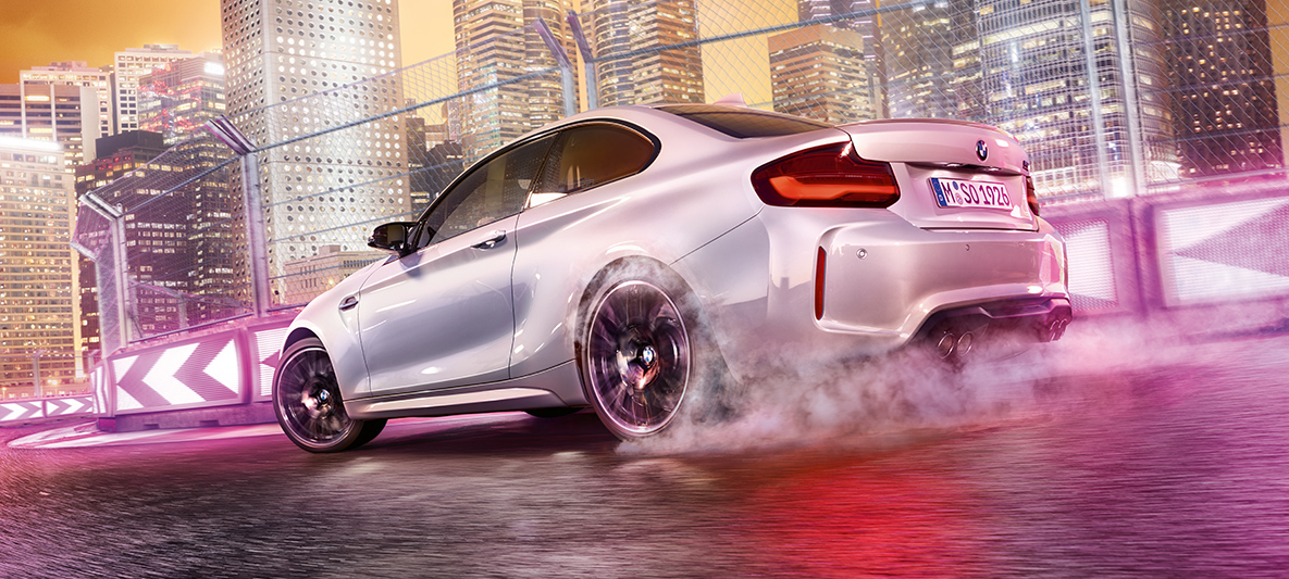La BMW M2 Competition accélère. Une BMW M2 Competition blanche driftant sr le circuit de course, devant une skyline nocturne.